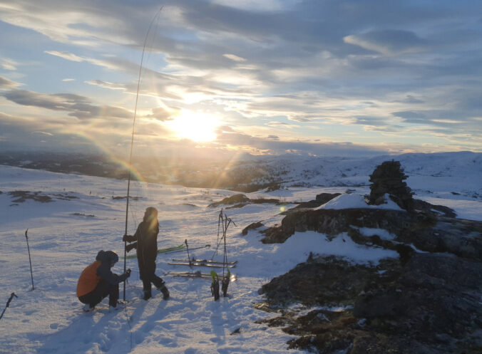 Two people are setting up an antenna in the snow. In the middle, there are several skis, a pair of snowshoes and ski poles in the snow. On the right, the summit is marked by a pile of stones. In the background, the low sun shines behind a few clouds.